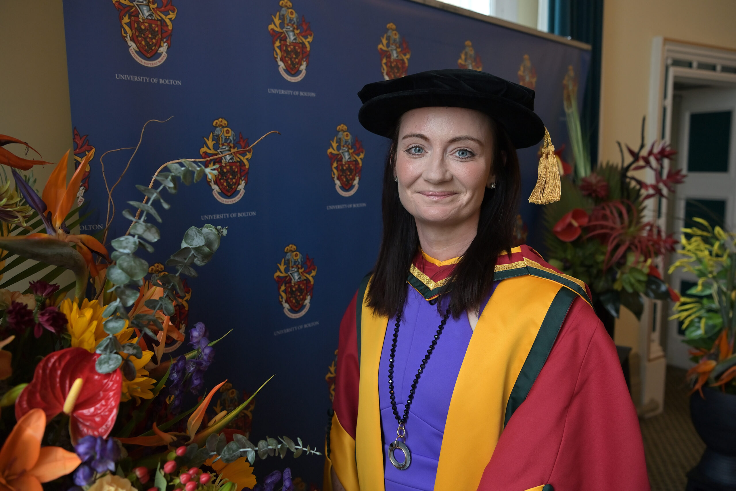 Dr Helen Wall in her graduation robes of red with a yellow collar and the black cap