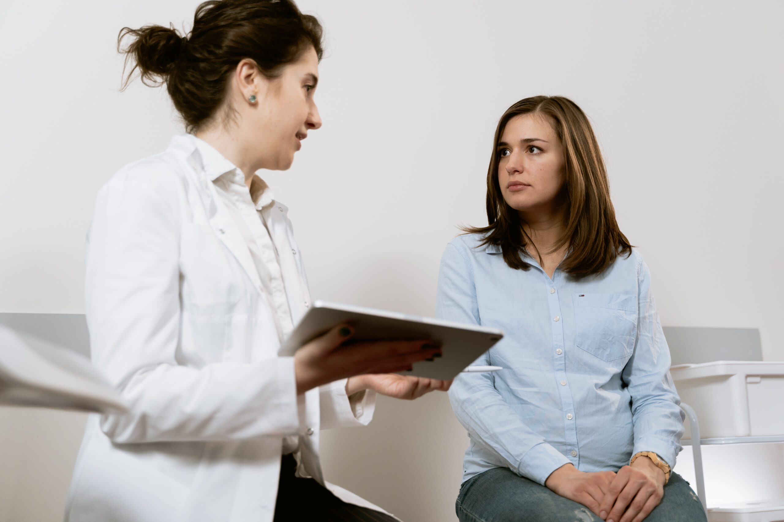 Salford innovation fund - A photo of a female staff member in a white coat holding a tablet computer next to a female patient in a blue shirt. They are looking at each other. The background is white.
