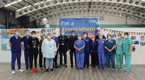 Mayor and staff in uniform against a backdrop of a blue sign with white and yellow text saying "I am a vaccine hero"