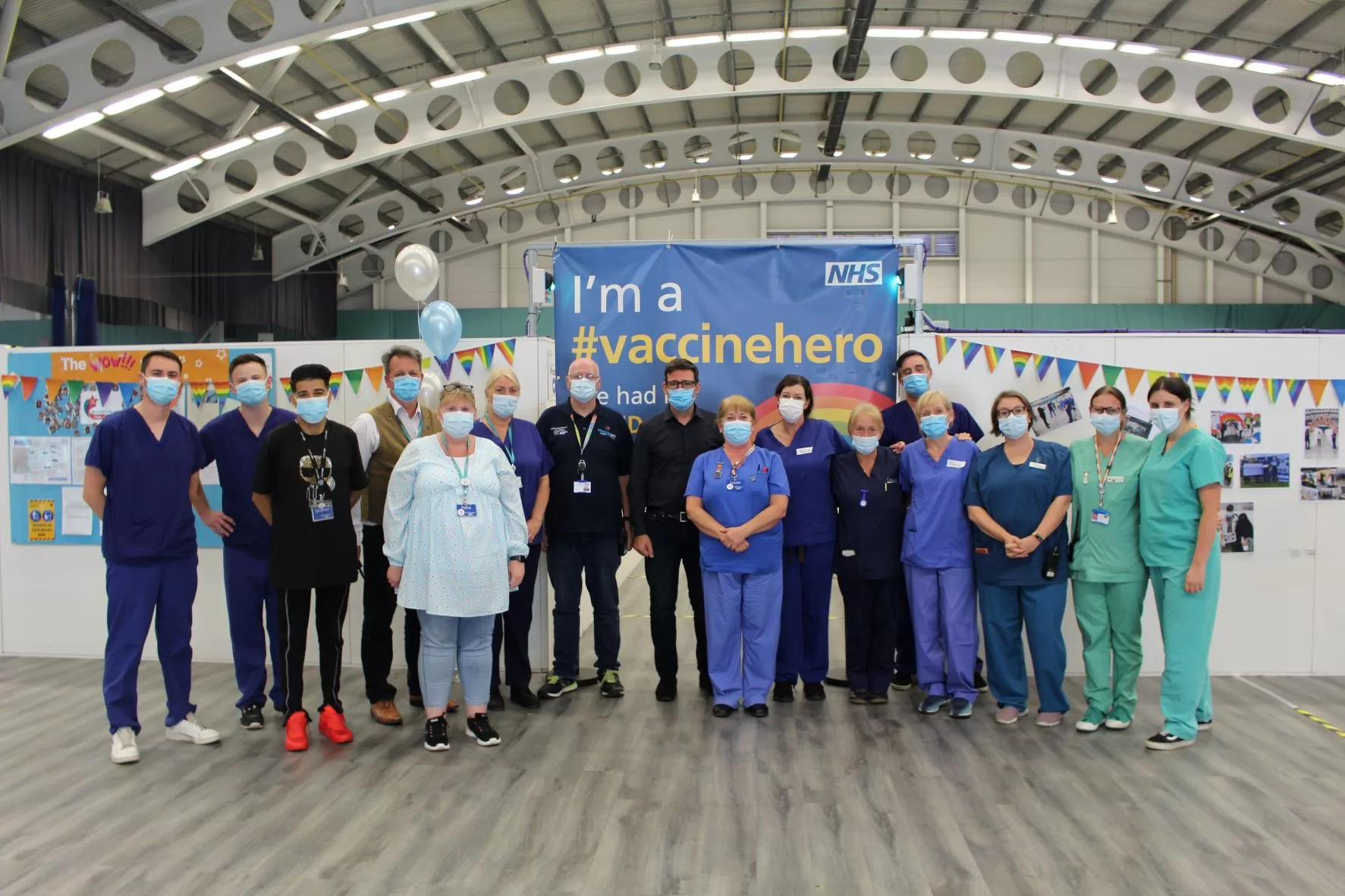Mayor and staff in uniform against a backdrop of a blue sign with white and yellow text saying "I am a vaccine hero"