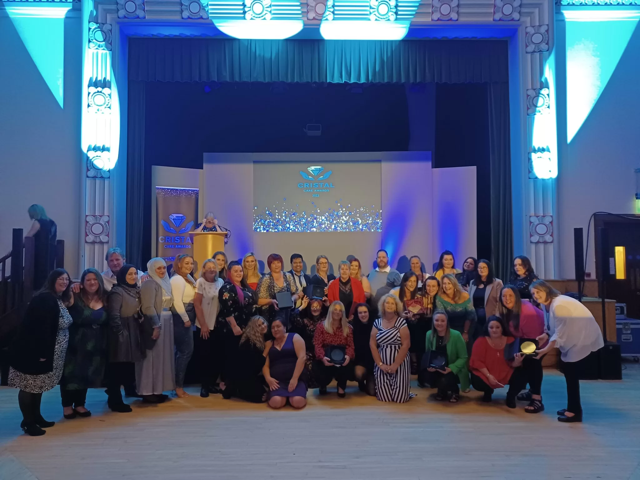 A group of care home and domiciliary care professionals, men and women, standing together in rows in front of a stage showing the CRISTAL Care Awards 2022 logo, holding their winning trophies.