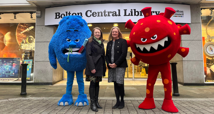 Two women wearing dresses and black coats standing together outside Bolton Central Library. They are in between a giant blue flu monster with a runny nose and a thermometer in its mouth and a giant red monster in the shape of a Covid virus.