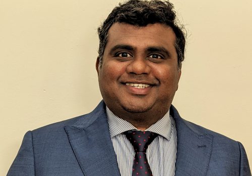 Dr Murugesan Raja wearing a light blue coloured suit jacket, with blue and white stripped shirt and a navy blue and red polkadot tie, standing in front of a cream background.