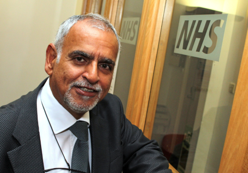 Former NHS Bolton Clinical Commissioning Group chairman Dr Wirin Bhatiani. He is wearing a suit and tie and leaning against a glass door with the NHS logo etched on it.