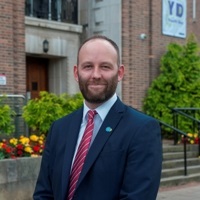 City Mayor Paul Dennett is standing outside in a red tie, light blue shirt and blue suit blazer. There are flowers and a building behind him.
