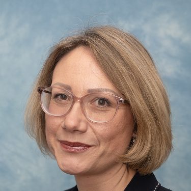 Lynne is chief executive of Bury Council. The photo shows her smiling at the camera. She wears glasses and is in a black jacket.