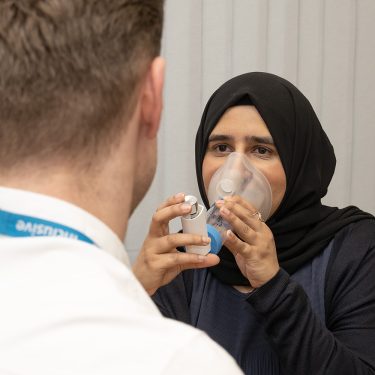 A person using an inhaler and spacer while a clinician watches.