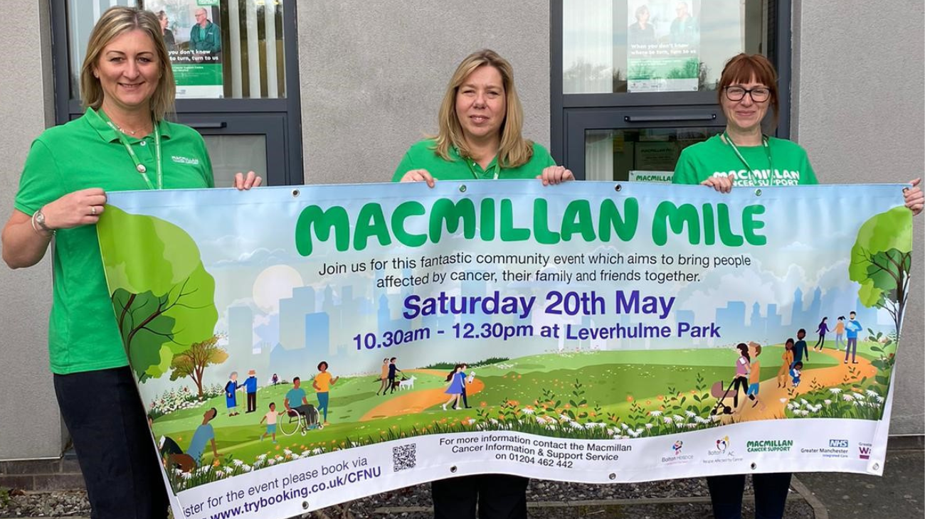 Three people wearing green polo shirts. They are holding up a banner advertising the Macmillan Mile walk which takes place in Bolton on May 20, 2023.