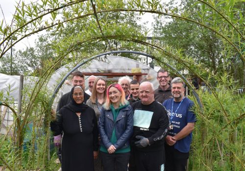 A group of nine smiling people standing in a tunnel made of bamboo growing over arched supports in the Petrus Community Garden.