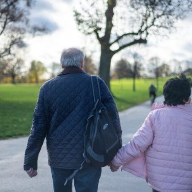 Two older people walking through a park shown from behind. They are holding hands. There are trees and grass.