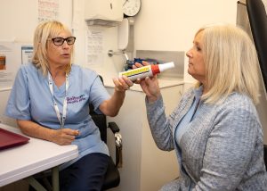 Practice nurse Sylvia sitting at a desk in a consultation room. She is talking to a patient as they use an inhaler. 