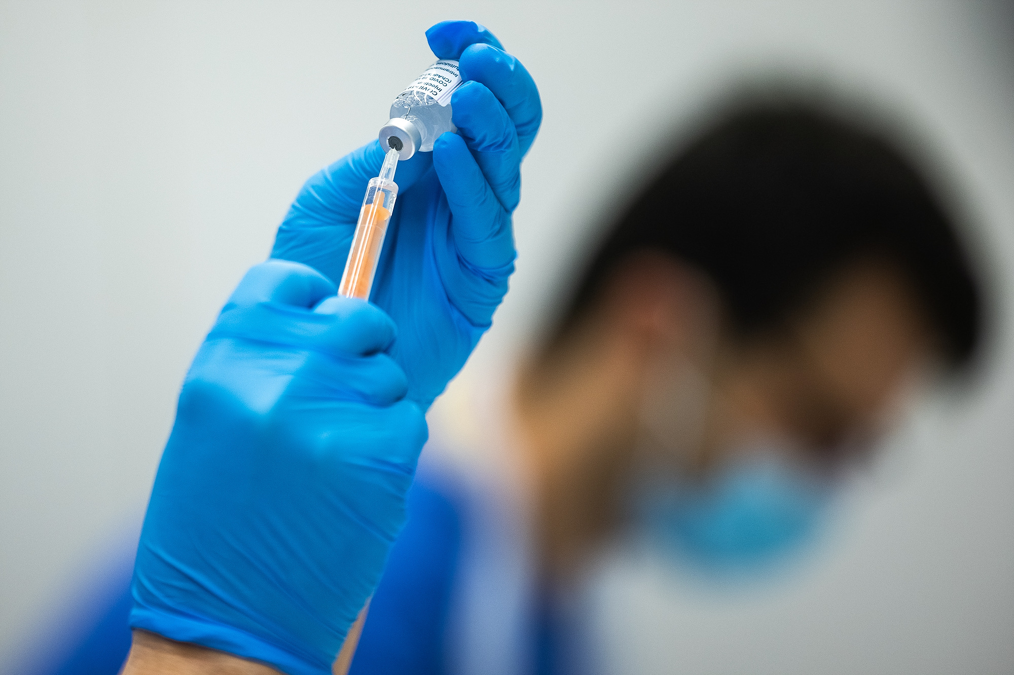 A pair of hands in blue gloves drawing up a vaccine in to a needle, with a blurred, masked person in scrubs in the background.