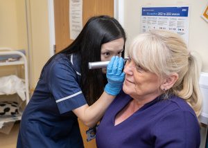 An advanced nurse practitioner wearing a blue uniform and examining a patient's ear with an otoscope