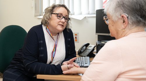 Care coordinator Cheryl sitting at a desk in a consulting room and talking to patient who is sitting opposite.