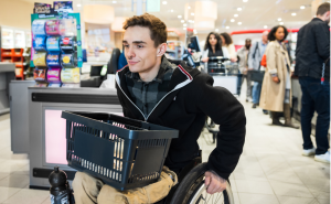 A wheelchair user in a supermarket with a shopping basket on their lap. They are wearing a striped polo shirt, a black hoodie, brown cargo pants and a nose ring. 