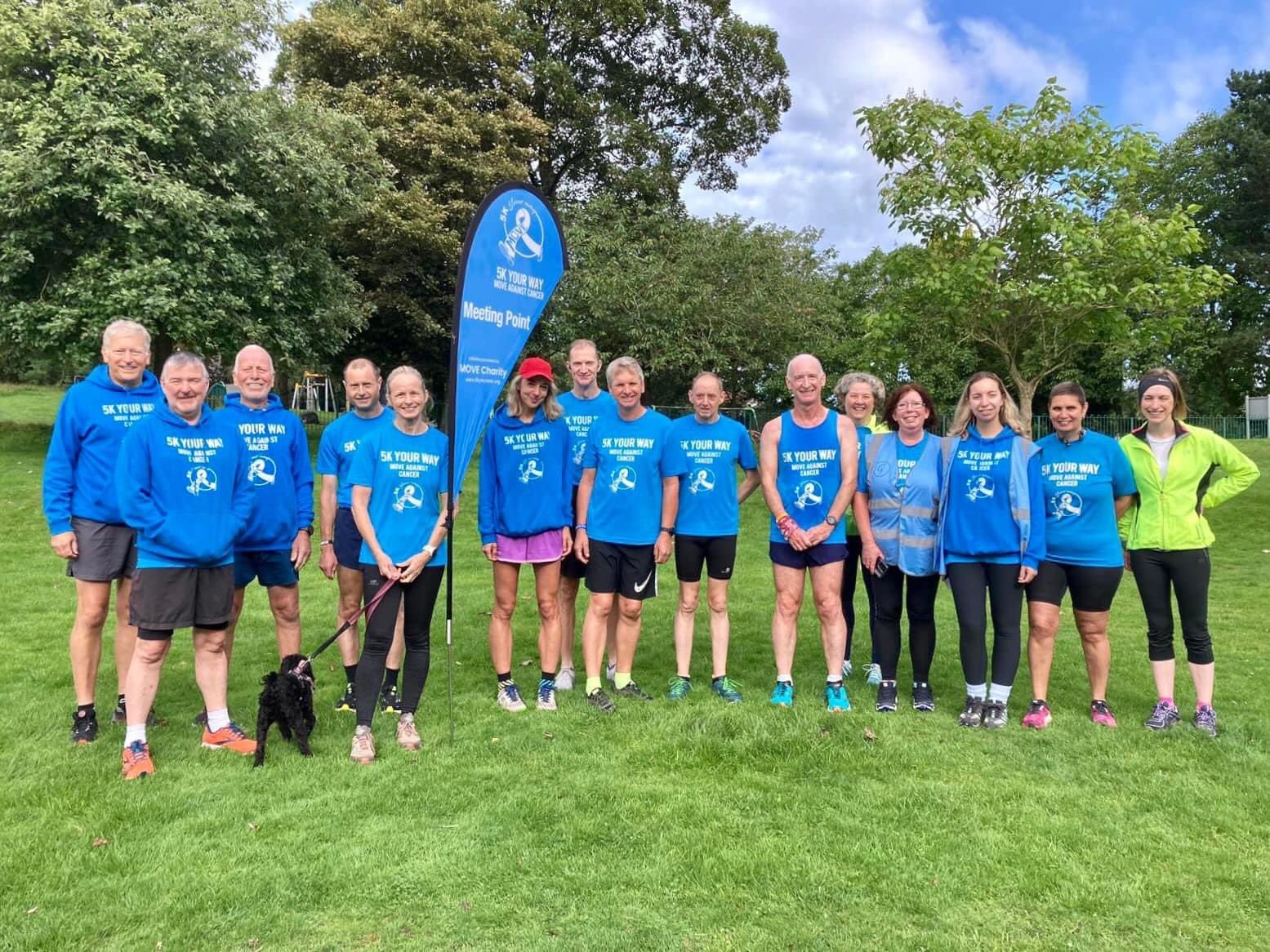 A large group of smiling people standing on grass in a park against a backdrop of green trees. They are wearing running and walking gear and blue tops with 5K Your Way on the front.