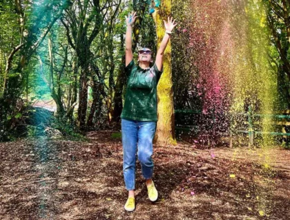 A person wearing jeans and a green polo shirt. They have a huge smile as they throw brightly-coloured powder in the air.