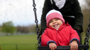 Child on a swing in a park. They are wearing a red coat and a pink wooly hat.