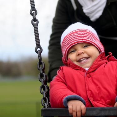 Child on a swing in a park. They are wearing a red coat and a pink wooly hat.