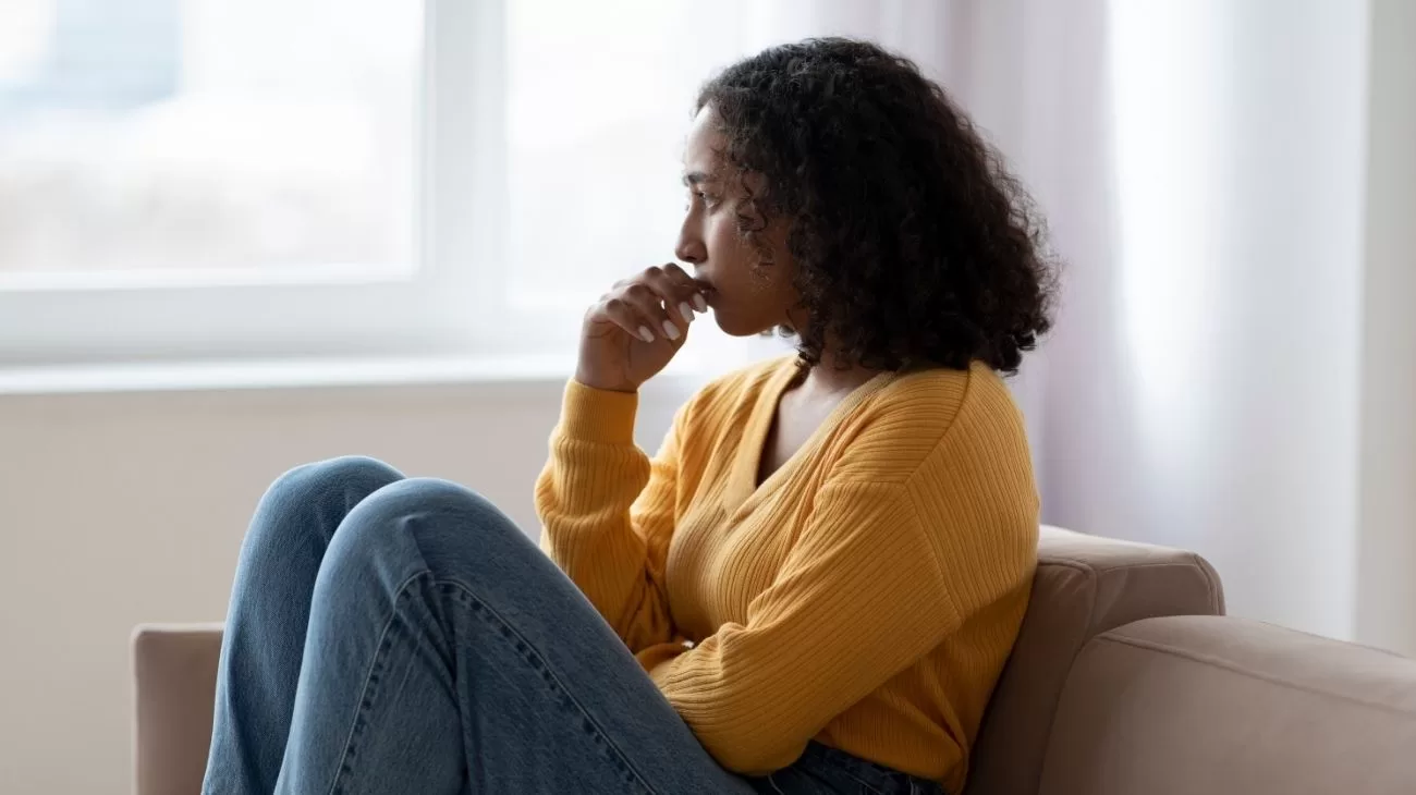 A woman sitting on a chair with her knees up in front of her. She is resting her chin on her hands and looking out of the window. The woman looks sad.