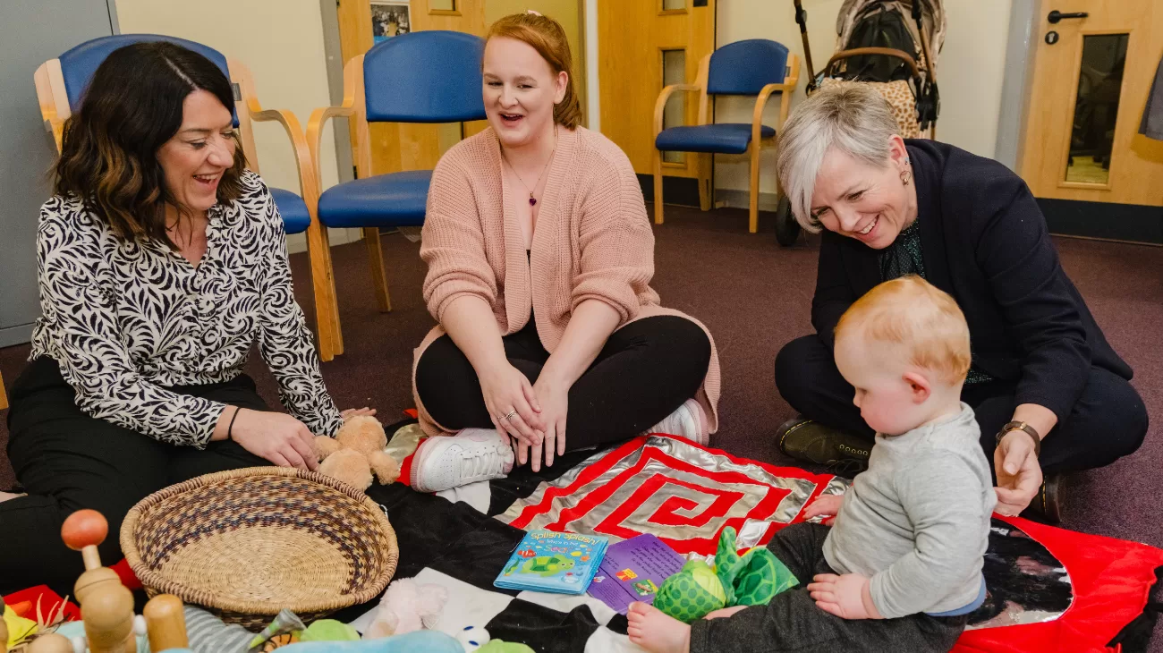 Three women are sat on the floor with a baby. The baby is looking at toys and sat on a play mat
