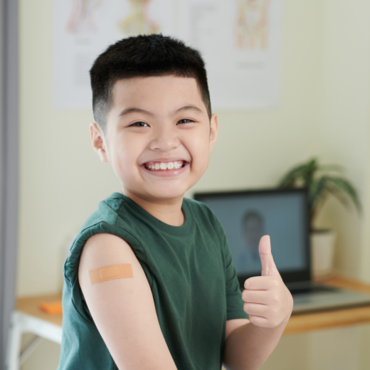 A young child smiling at the camera with their thumb up. Their t-shirt sleeve has been rolled up and there is a plaster at the top of his arm where they have had an injection. You can see a laptop and a desk in the background.