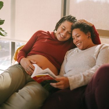 A pregnant person sat on the sofa with another person, looking at a book and smiling.