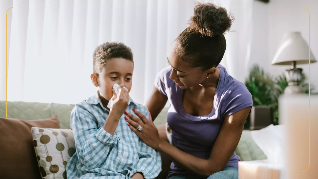 A mum and son sat on a sofa in a living room. The child is wearing pyjamas and his blowing his nose in to a tissue. His mum has her hand on his shoulder supportively.