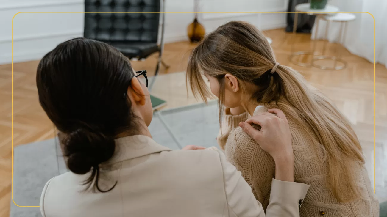 Two women are sat on a sofa with their back to the camera. One woman looks upset, the other woman has her hand on her shoulder in a supportive way. They are sat in an office.