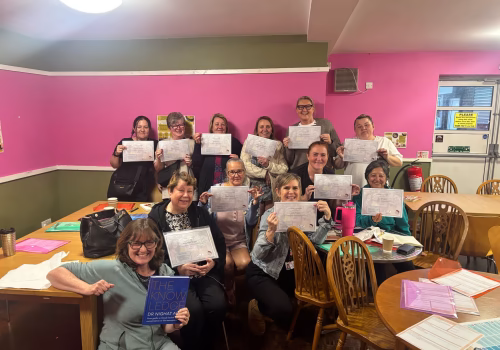Group photo of twelve individuals at the North Manchester Menopause Hub, posed in a classroom setting with pink and green walls. The front row is seated and the back row is standing, with several people holding certificates and one person displaying a book titled 'THE KNOWLEDGE' by Dr. Khayat. Tables in the room have papers, folders, and drinks, suggesting a celebratory or educational event."