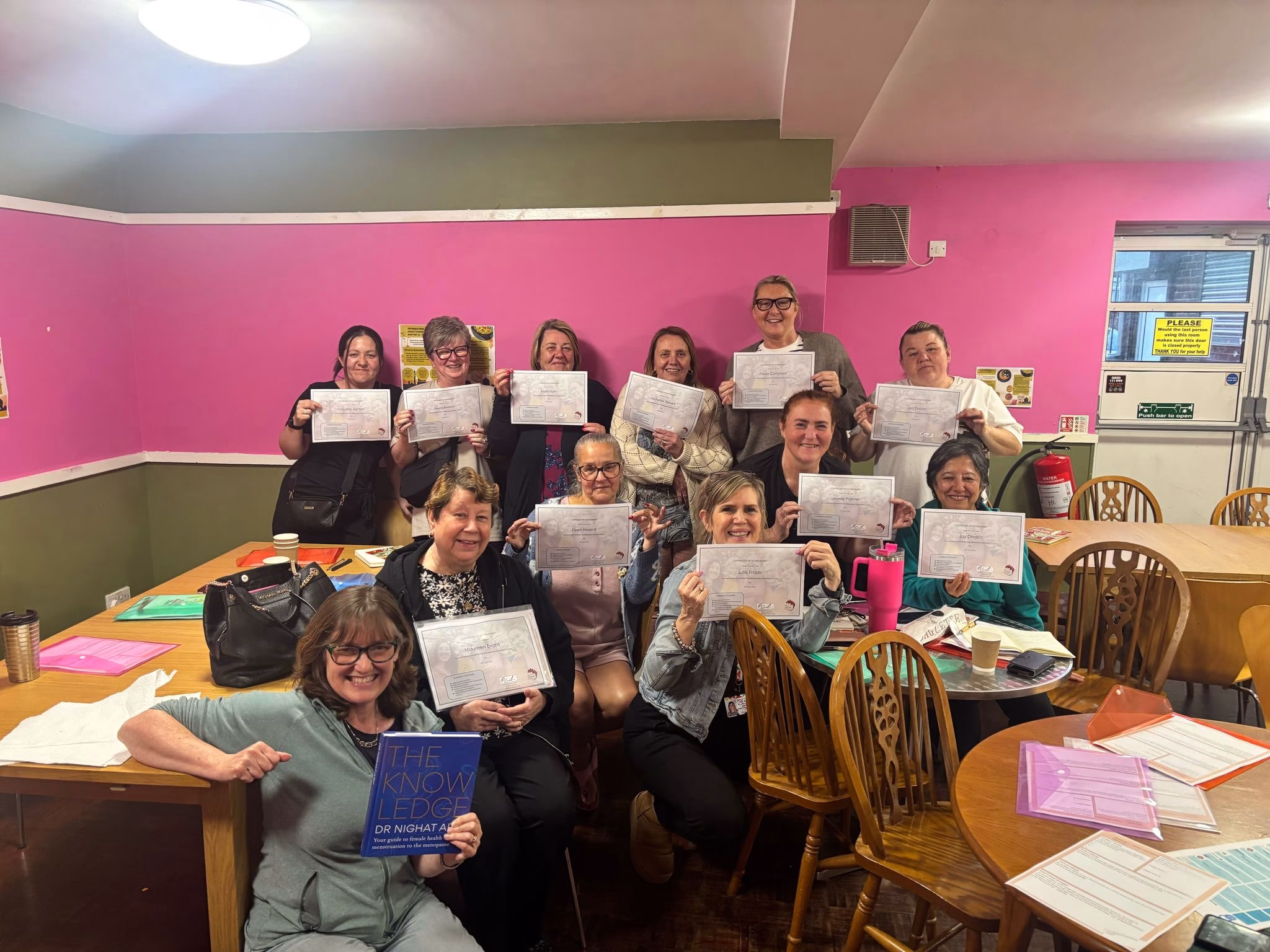 Group photo of twelve individuals at the North Manchester Menopause Hub, posed in a classroom setting with pink and green walls. The front row is seated and the back row is standing, with several people holding certificates and one person displaying a book titled 'THE KNOWLEDGE' by Dr. Khayat. Tables in the room have papers, folders, and drinks, suggesting a celebratory or educational event."