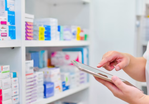 A person stood holding a device with pharmacy shelves in the background.