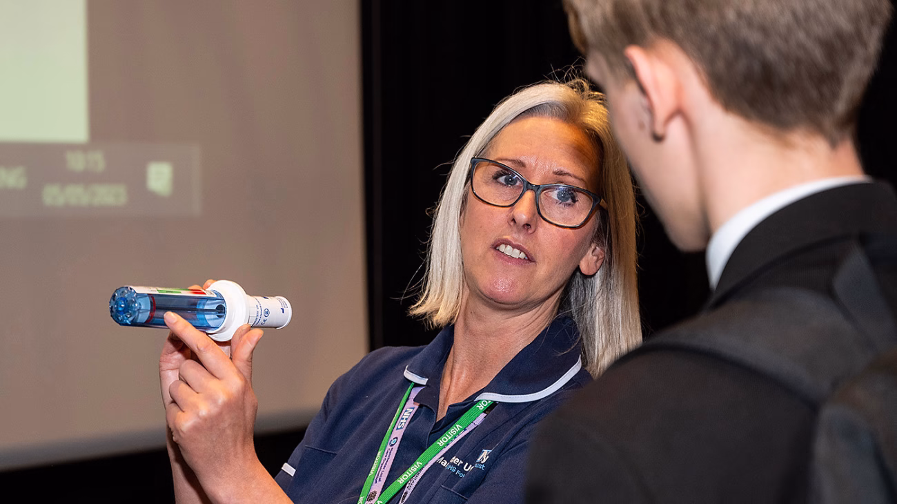 Head and shoulders shot of a school nurse wearing a dark blue uniform holding an asthma inhaler and spacer as a pupil looks on.