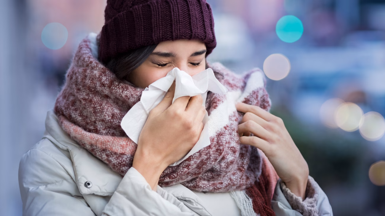 Person in a beanie, scarf and coat sneezing into a tissue